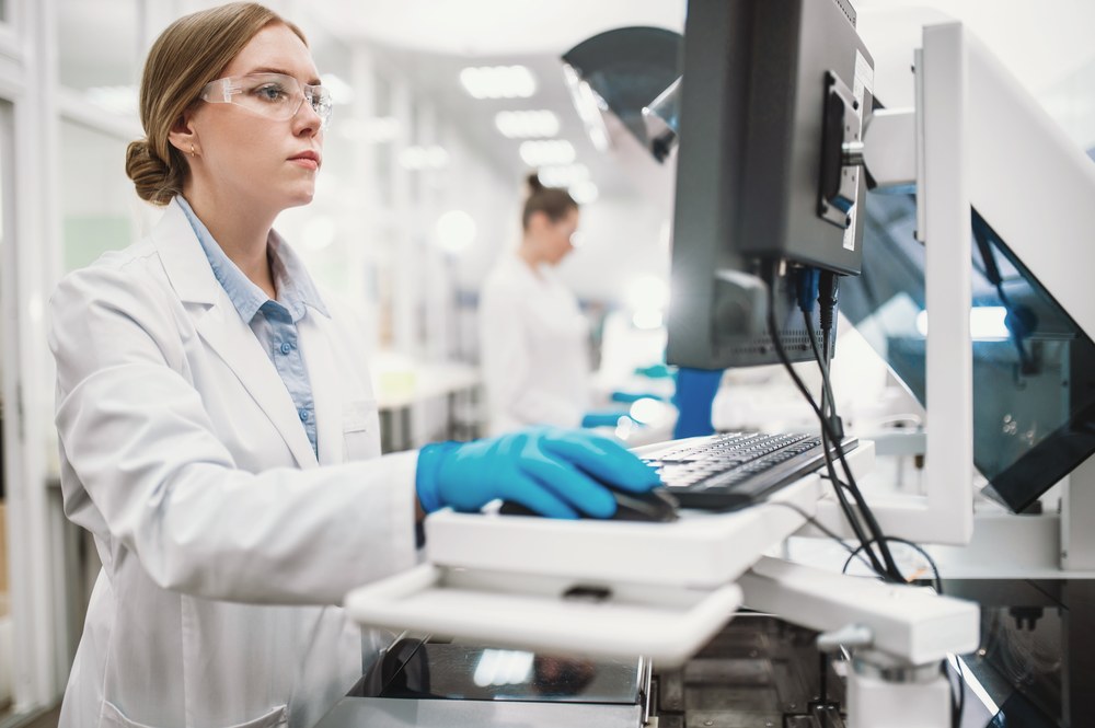 A researcher in a lab coat and safety glasses operating an analytical instrument in a modern laboratory.